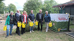 Menschen stehen mit Gießkannen neben einem Regenwasserfass