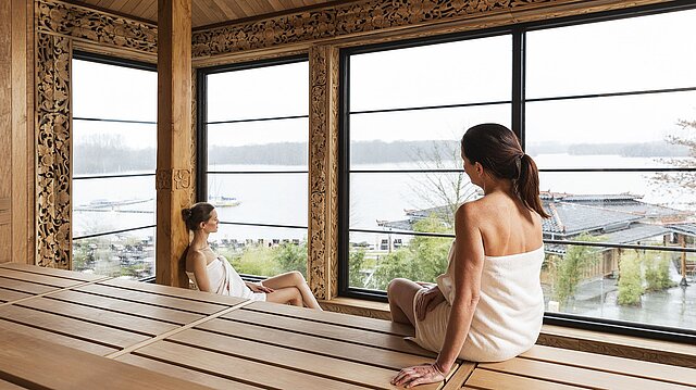 Zwei Frauen sitzen im Inneren einer Sauna vor einer Glasfront mit Blick auf den Elbsee.