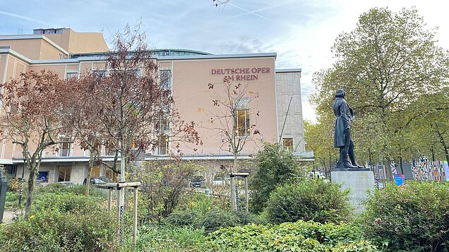 Die Deutsche Oper von der Parkseite aus mit viel Grün und einer Statue im Vordergrund.